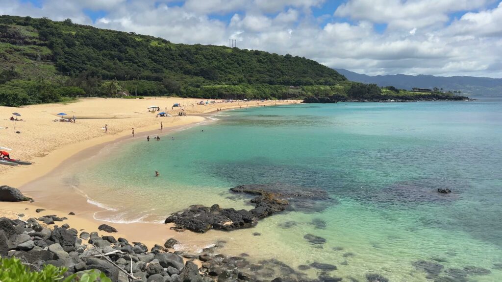 Calm turquoise water, sandy shoreline, and black lava rocks at Waimea Bay on Oahu.