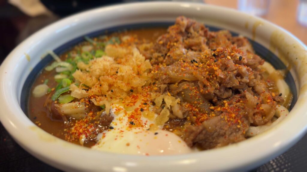 Spicy curry nikutama udon topped with beef, green onions, tempura flakes, chili seasoning, and a soft-cooked egg.