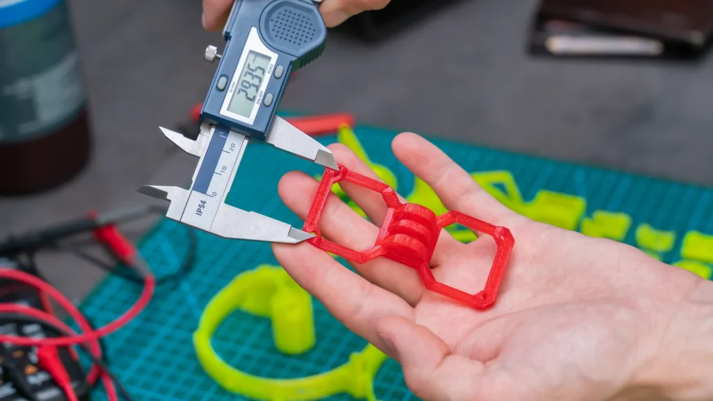 A person holds a red 3D printed part while measuring an edge with a digital caliper.