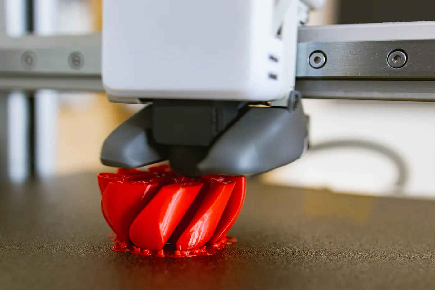 A filament printer head is positioned above a partially printed red object with curved grooves.