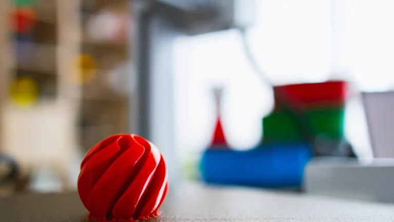 A red grooved spherical object sits on a printer bed with other printed shapes blurred behind it.