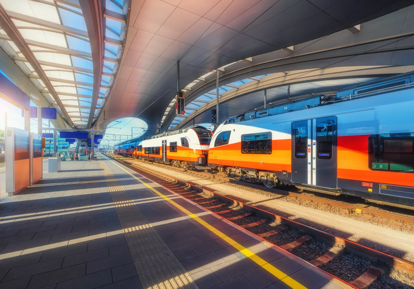 White, yellow, and orange passenger trains stand beside a covered railway platform.