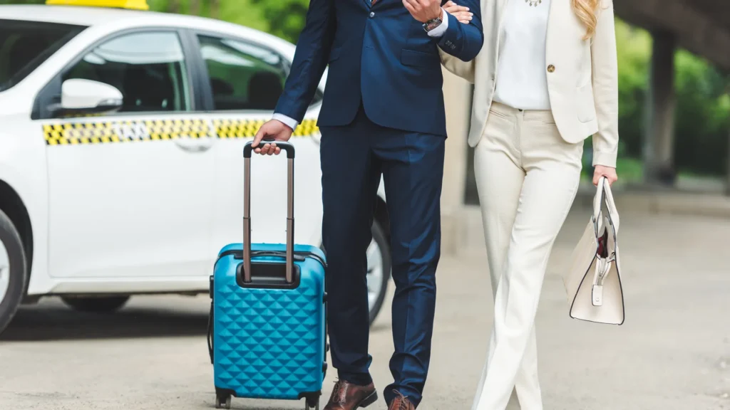 Two travelers walk with a blue wheeled suitcase beside a white taxi.