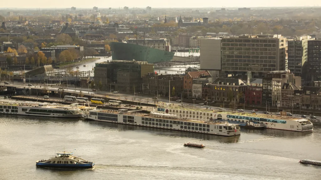 Blue and white ferry crossing the IJ in front of docked passenger vessels and Amsterdam buildings