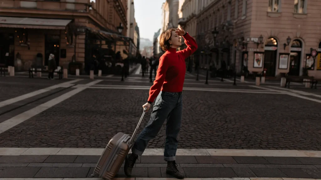 A traveler in a red sweater stands on a city street beside a rolling suitcase.