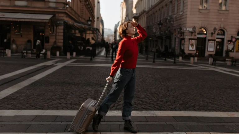 A traveler in a red sweater stands on a city street beside a rolling suitcase.