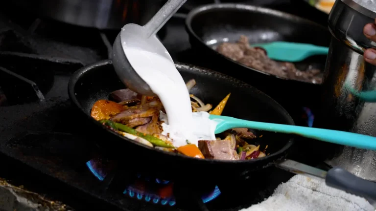A ladle pours white cream into a pan of ingredients on a stove.
