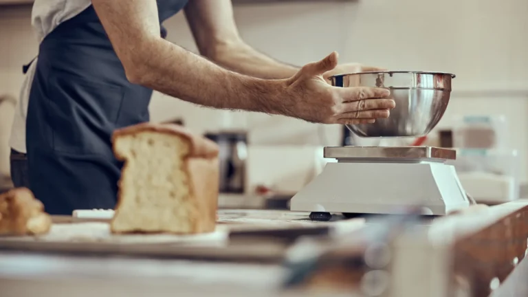 A baker holding a metal mixing bowl on a kitchen scale, with bread on the work surface.