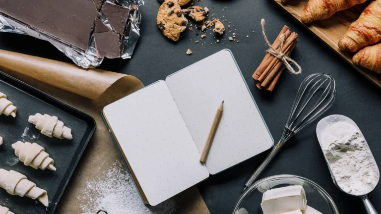 Open blank notebook and pencil surrounded by flour, butter, a whisk, dough, and pastries on a dark work surface