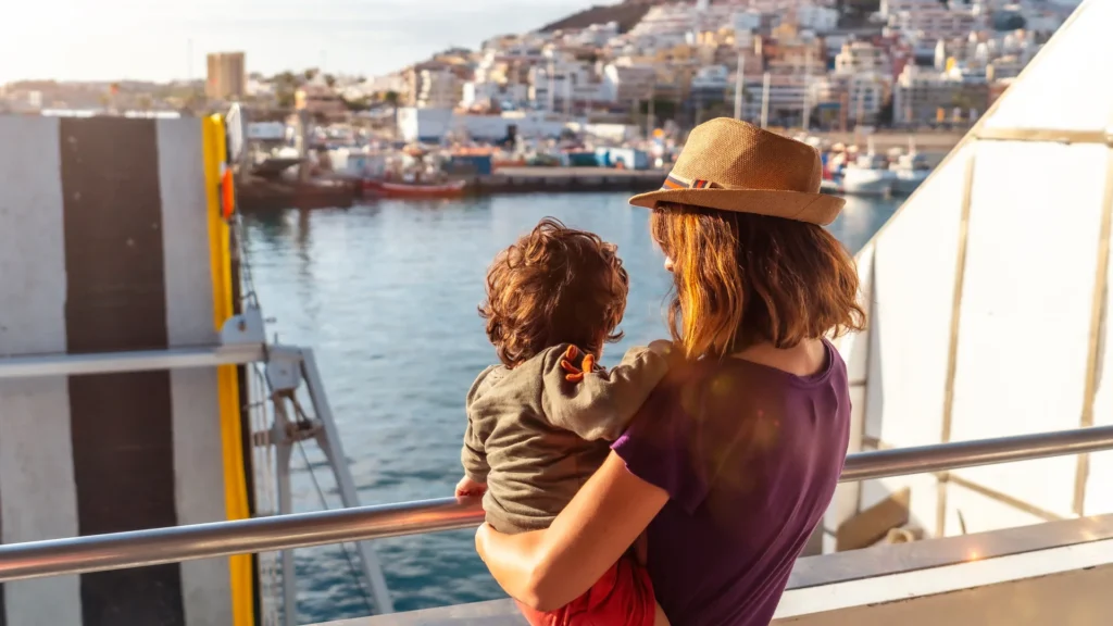 A parent holds a child at a boat railing while looking toward a coastal town.