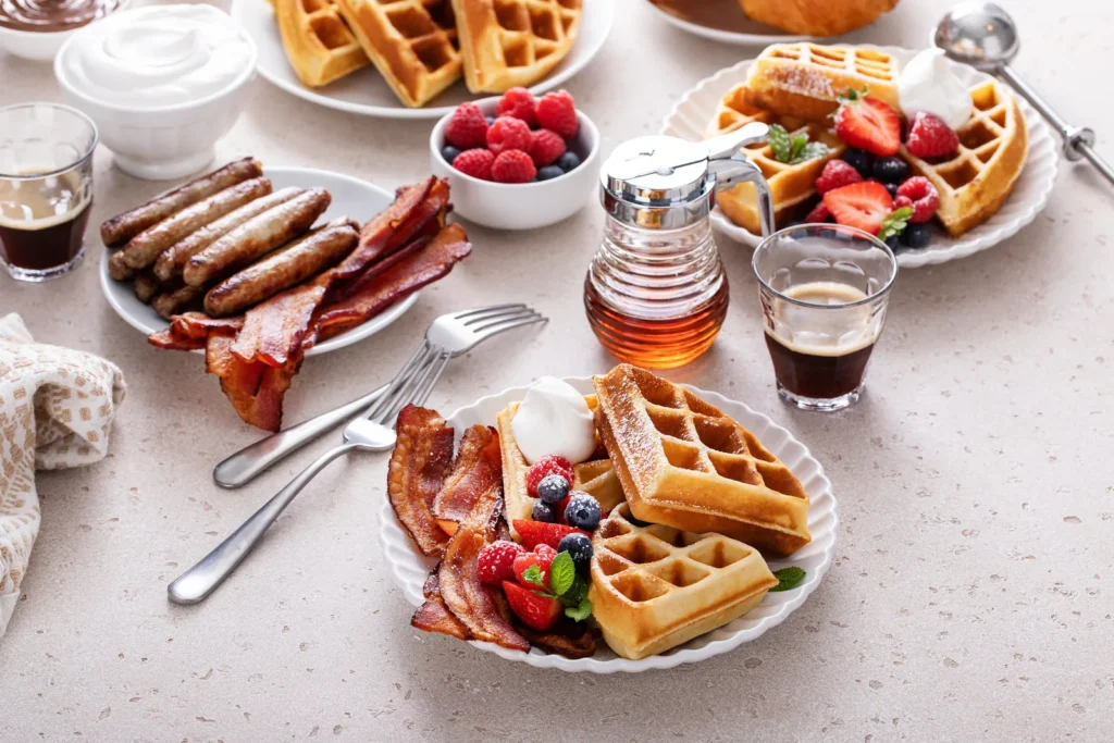 Waffles, berries, bacon, sausages, coffee, and syrup arranged across a light breakfast table.