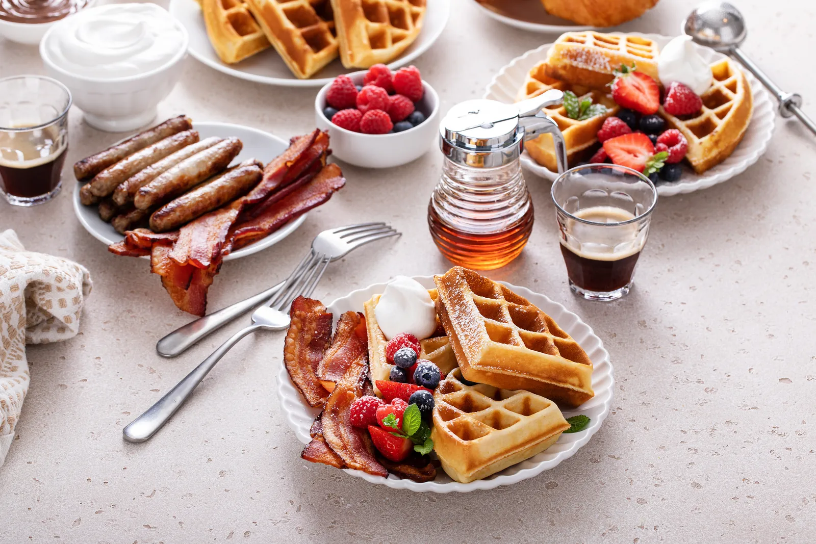 Waffles, berries, bacon, sausages, coffee, and syrup arranged across a light breakfast table.