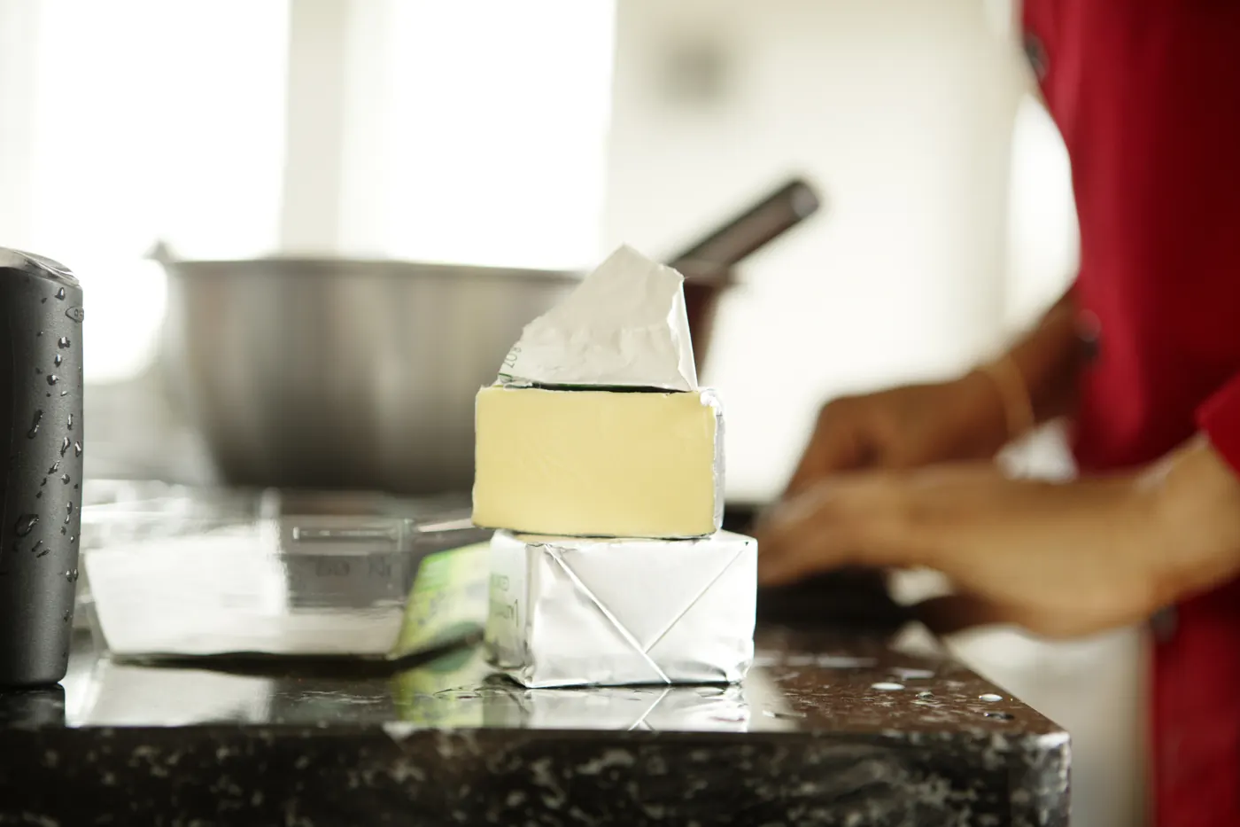 Partly unwrapped butter blocks on a kitchen counter with a mixing bowl and a person preparing dough in the background