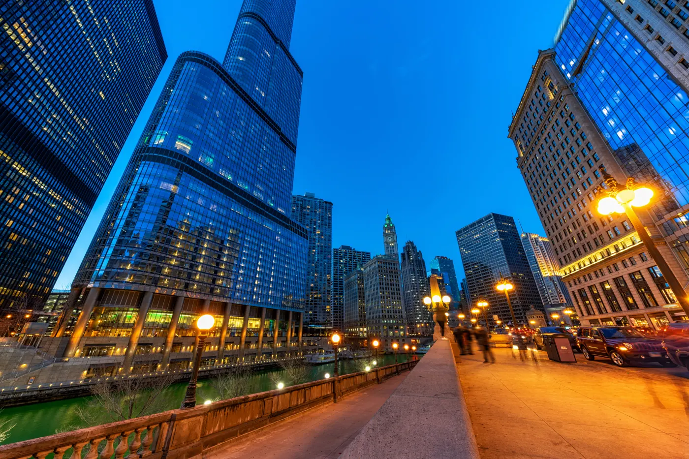 Riverfront sidewalk beside the Chicago River, with illuminated glass buildings and street traffic at blue hour