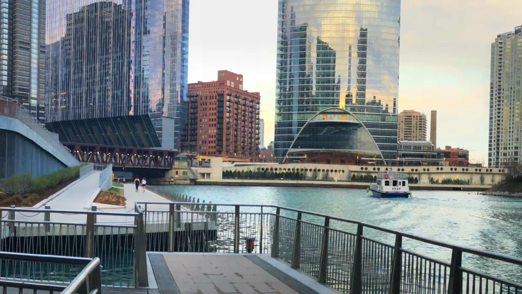 Chicago Riverwalk railings and paths beside the river, with a white boat and downtown buildings