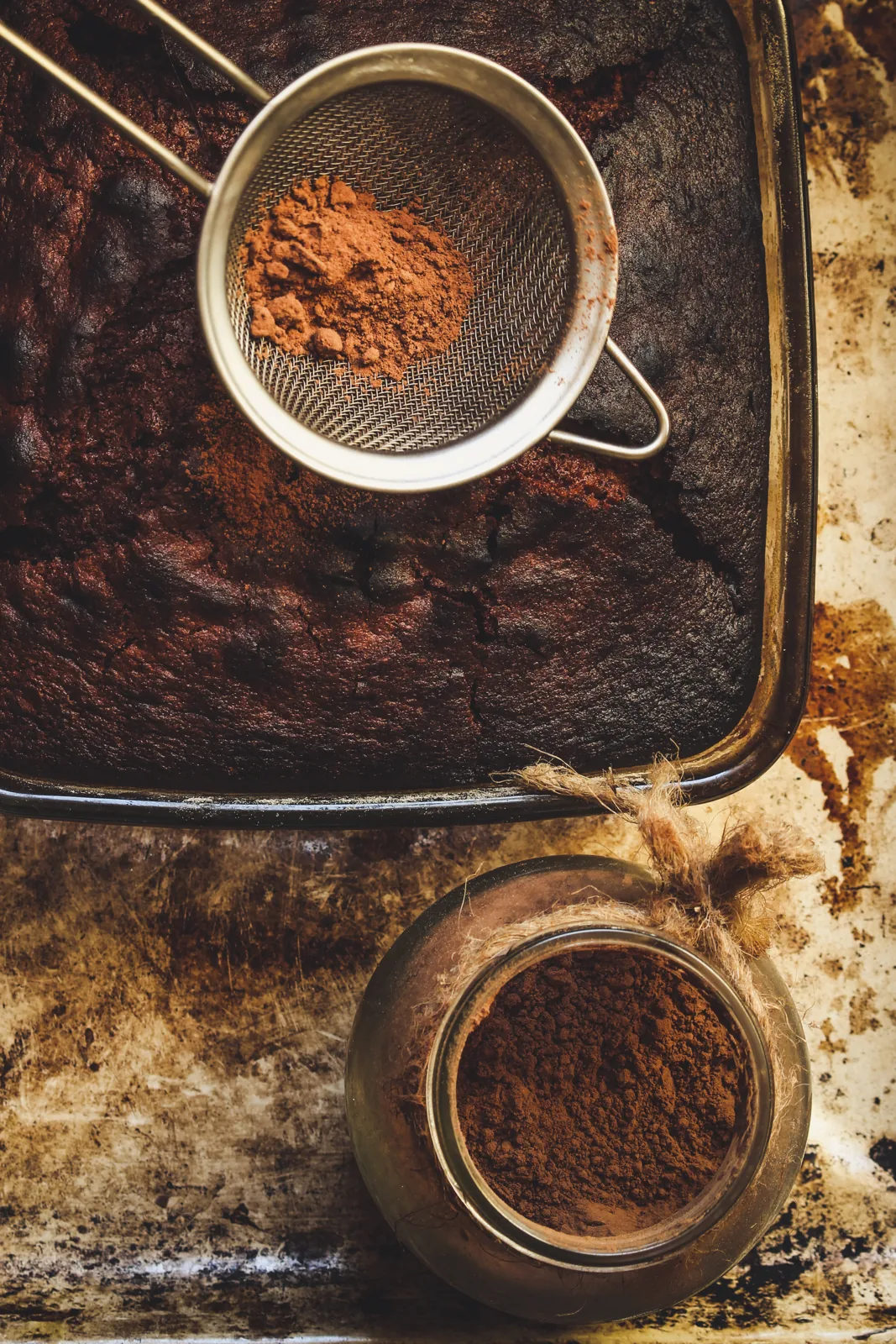 A tray of chocolate brownies beside a jar and sieve of cocoa powder.