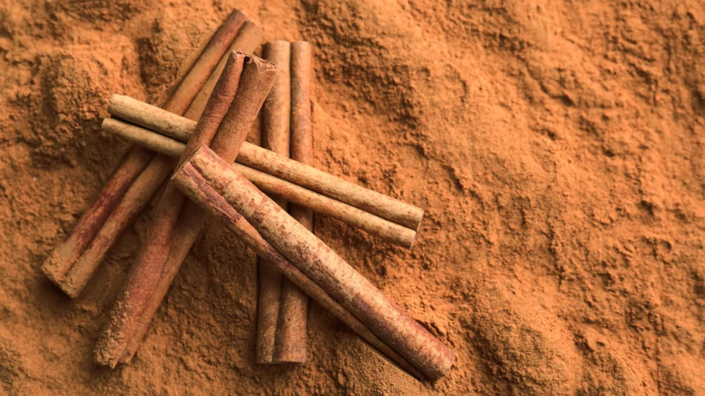 Several cinnamon sticks lie across a textured layer of ground cinnamon.