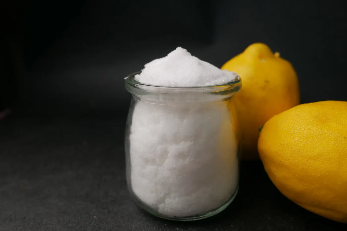 A glass jar of fine white citric acid stands beside two yellow lemons on a dark surface.