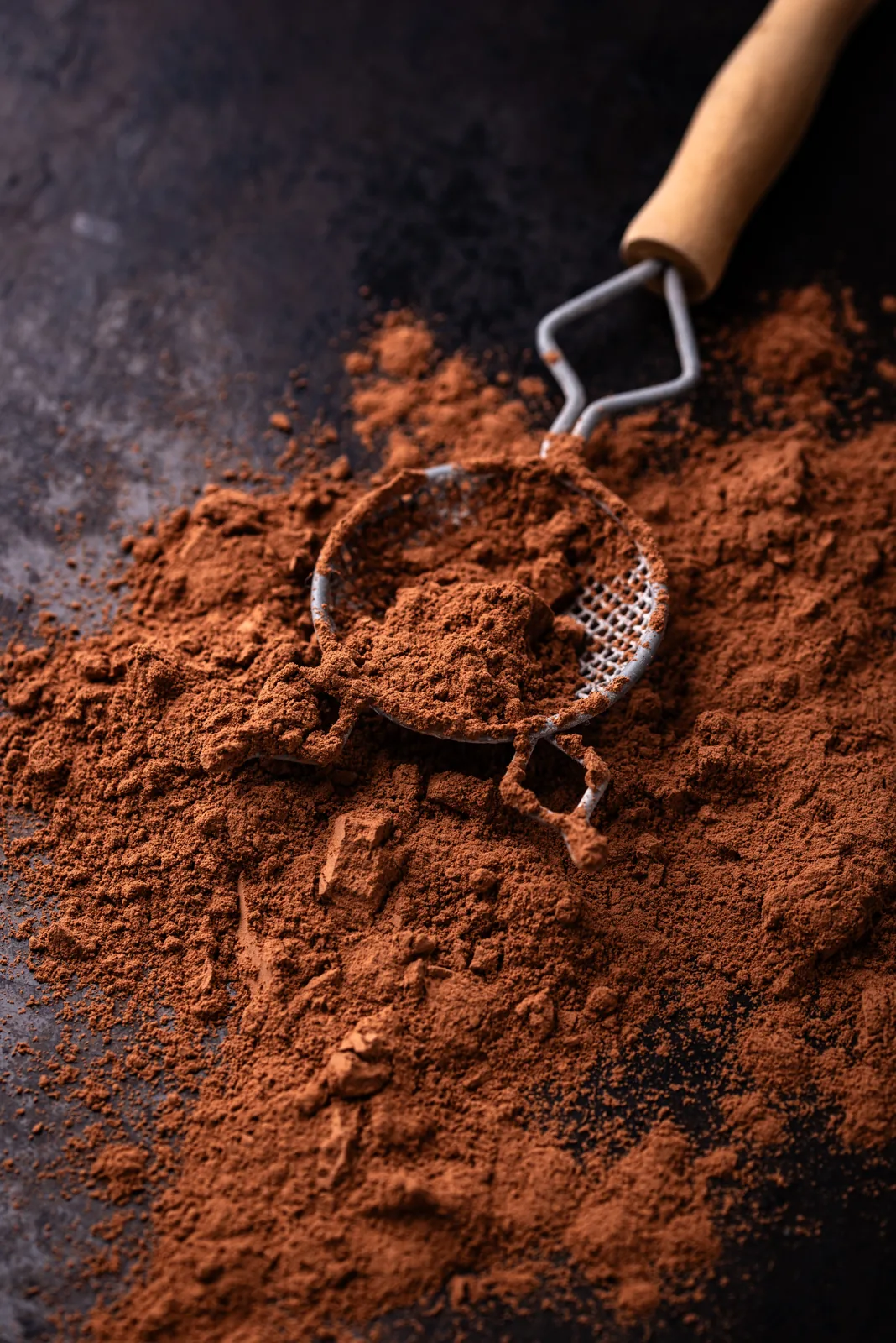 Cocoa powder scattered around a small metal sifter on a dark surface.