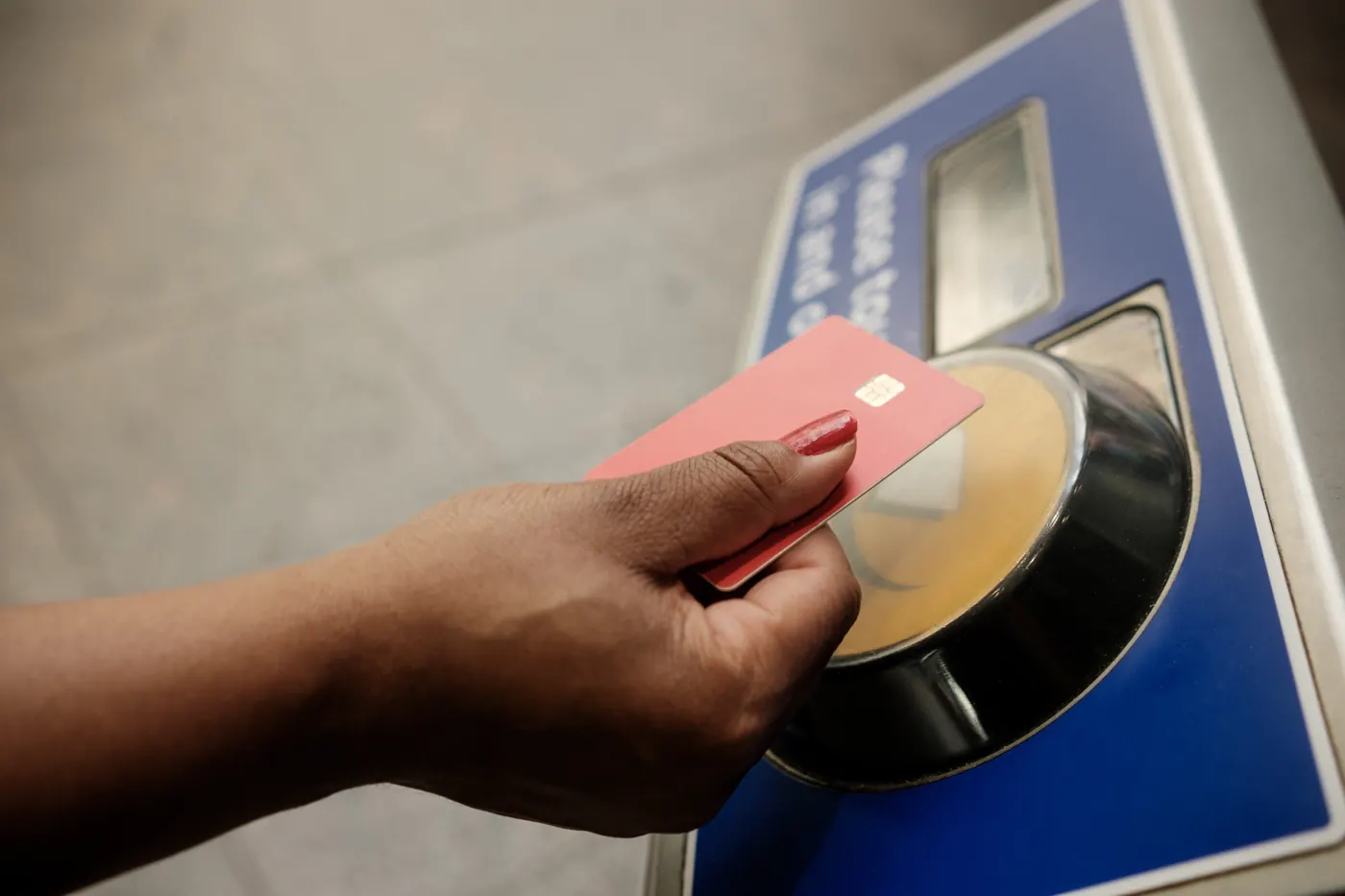 A hand holds a red payment card against a round yellow reader on a blue transit terminal.