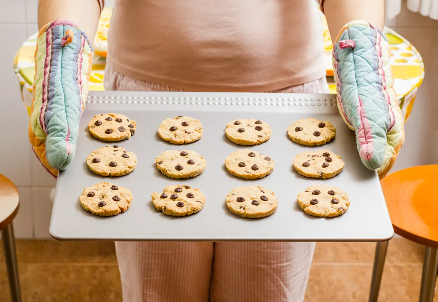 A person wearing oven mitts holding a metal baking sheet with rows of chocolate chip cookies