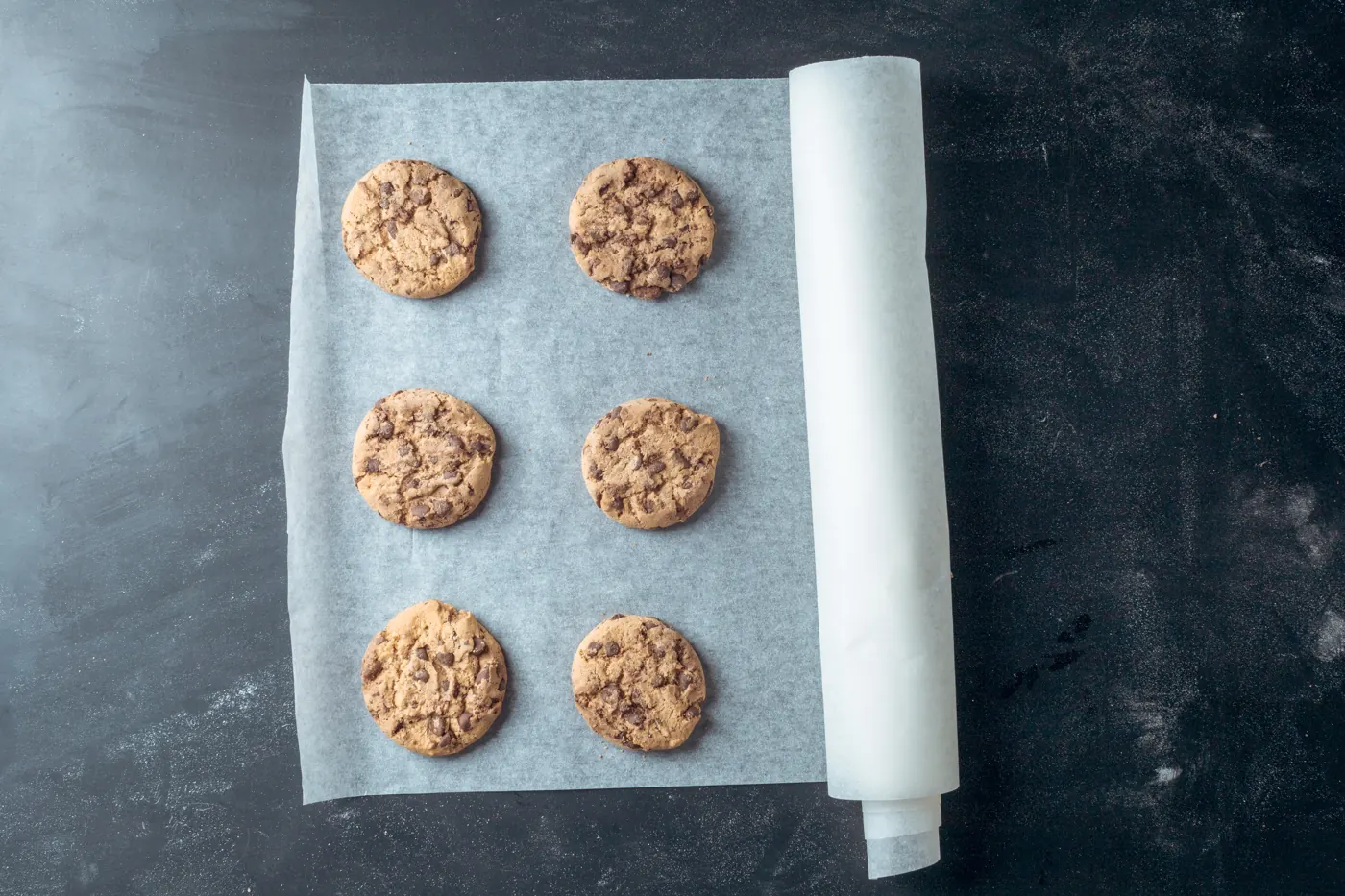 Six chocolate chip cookies resting on white parchment paper beside a partly unrolled sheet on a dark counter