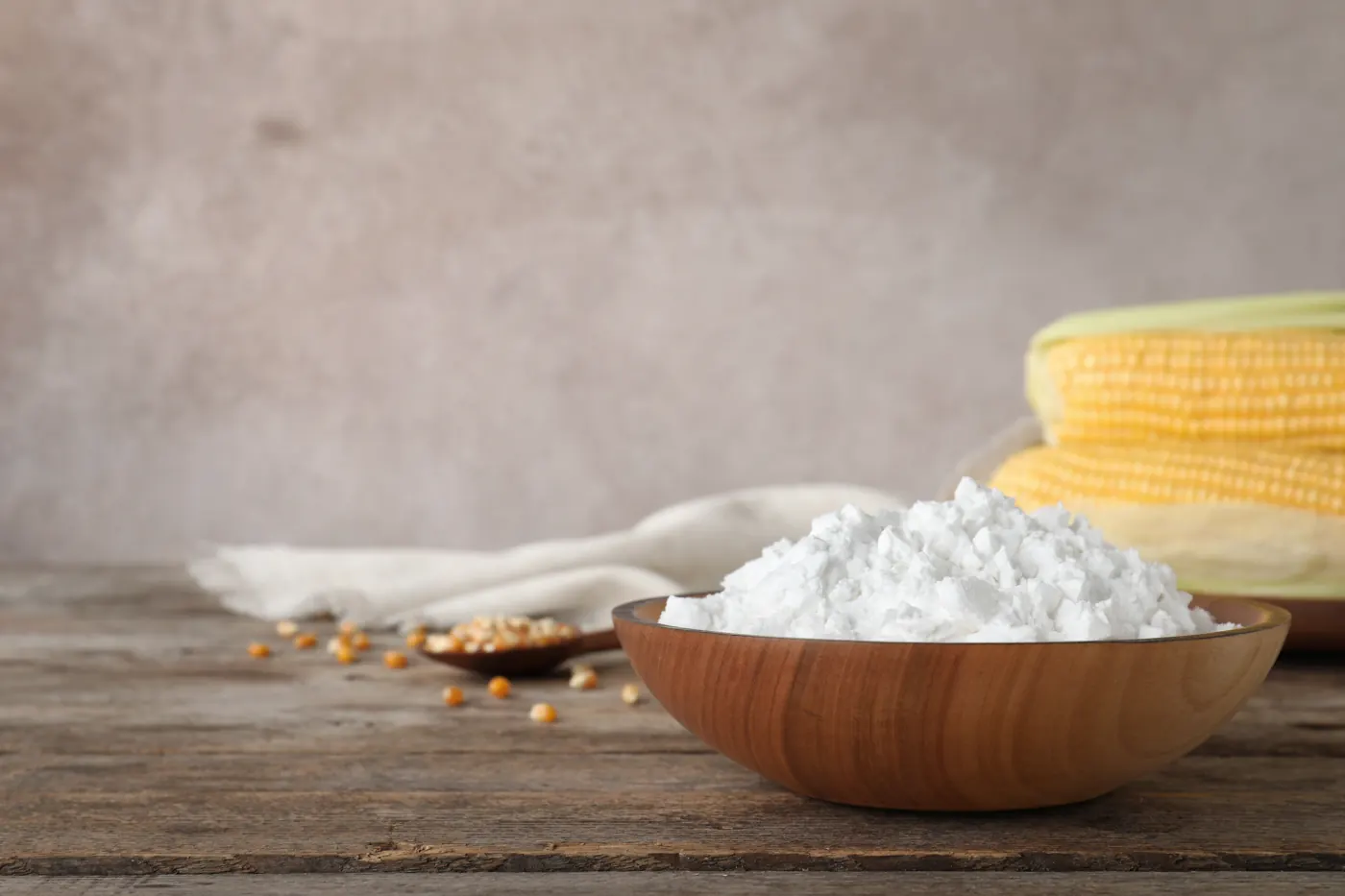 A wooden bowl holds white cornstarch beside corn cobs and scattered kernels.