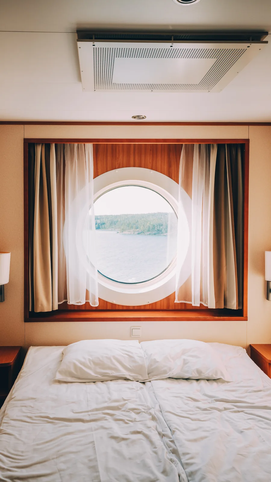 A bed, curtains, and a round porthole inside a compact ship cabin.