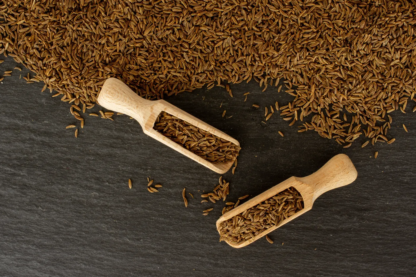 Whole cumin seeds lie with two small wooden scoops on a dark surface.