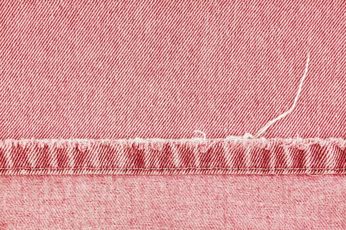Close-up of pink-red denim with a horizontal stitching line, frayed fabric edge, and loose white thread