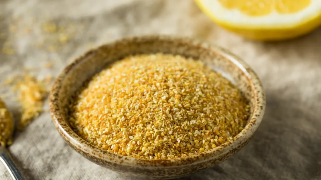 A small ceramic bowl holds finely dried yellow lemon peel, with a cut lemon behind it.