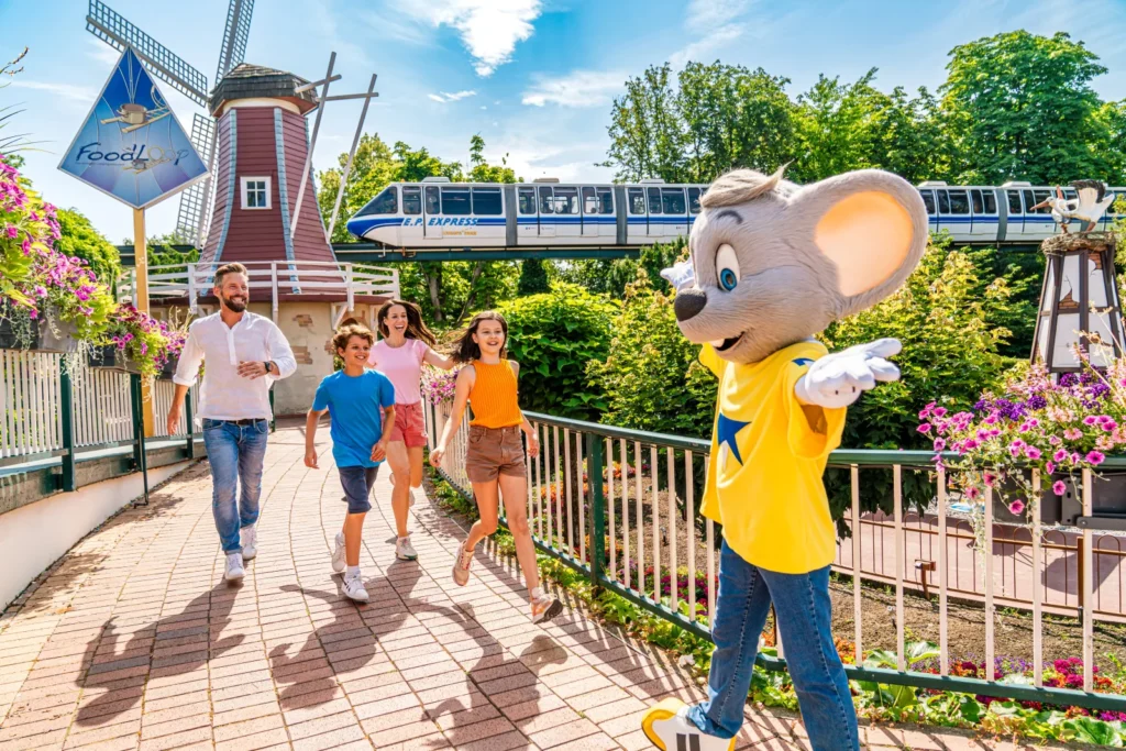 A family walking beside Ed Euromaus, a windmill, flowers, and an elevated train at Europa-Park.