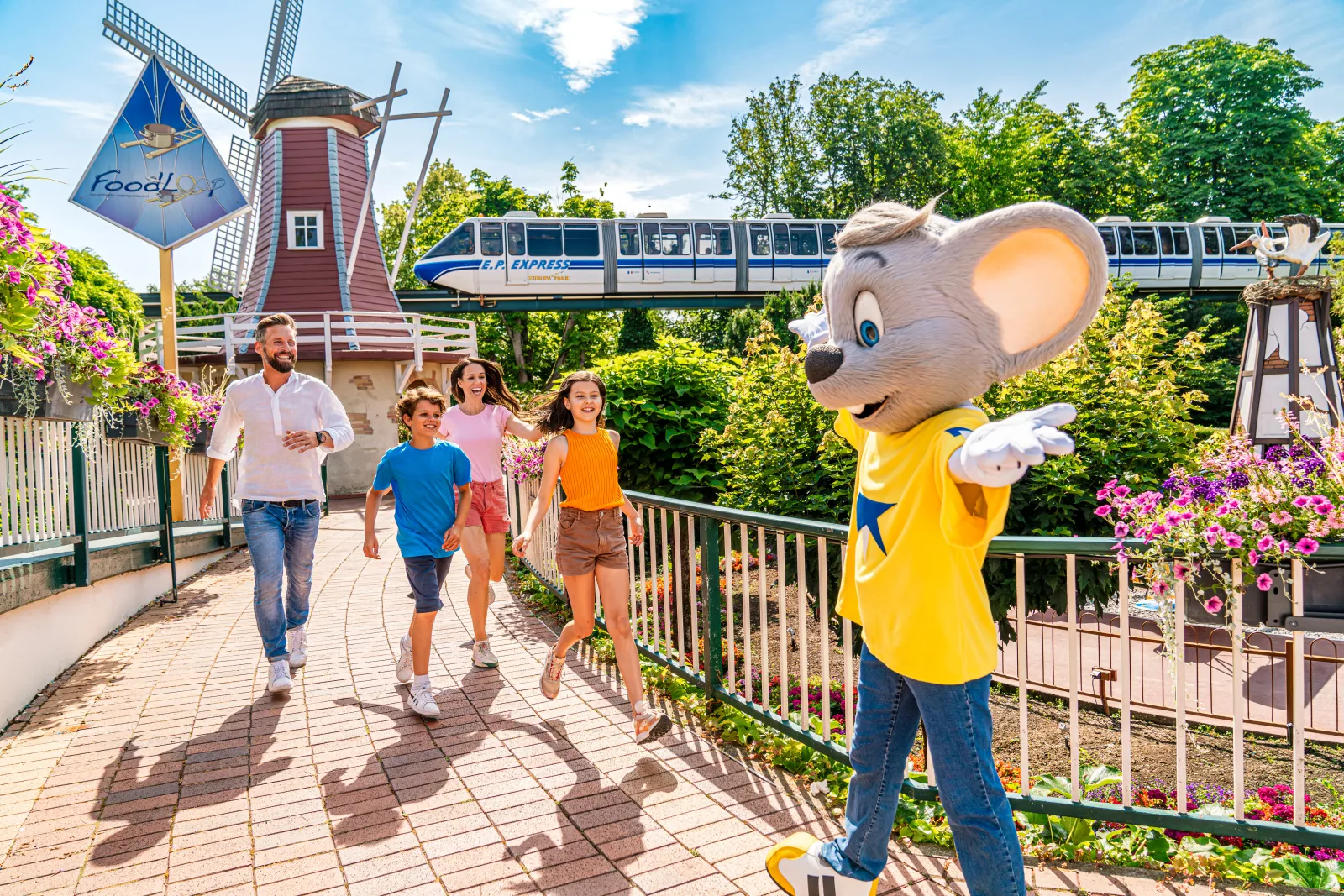 A family walking beside Ed Euromaus, a windmill, flowers, and an elevated train at Europa-Park.