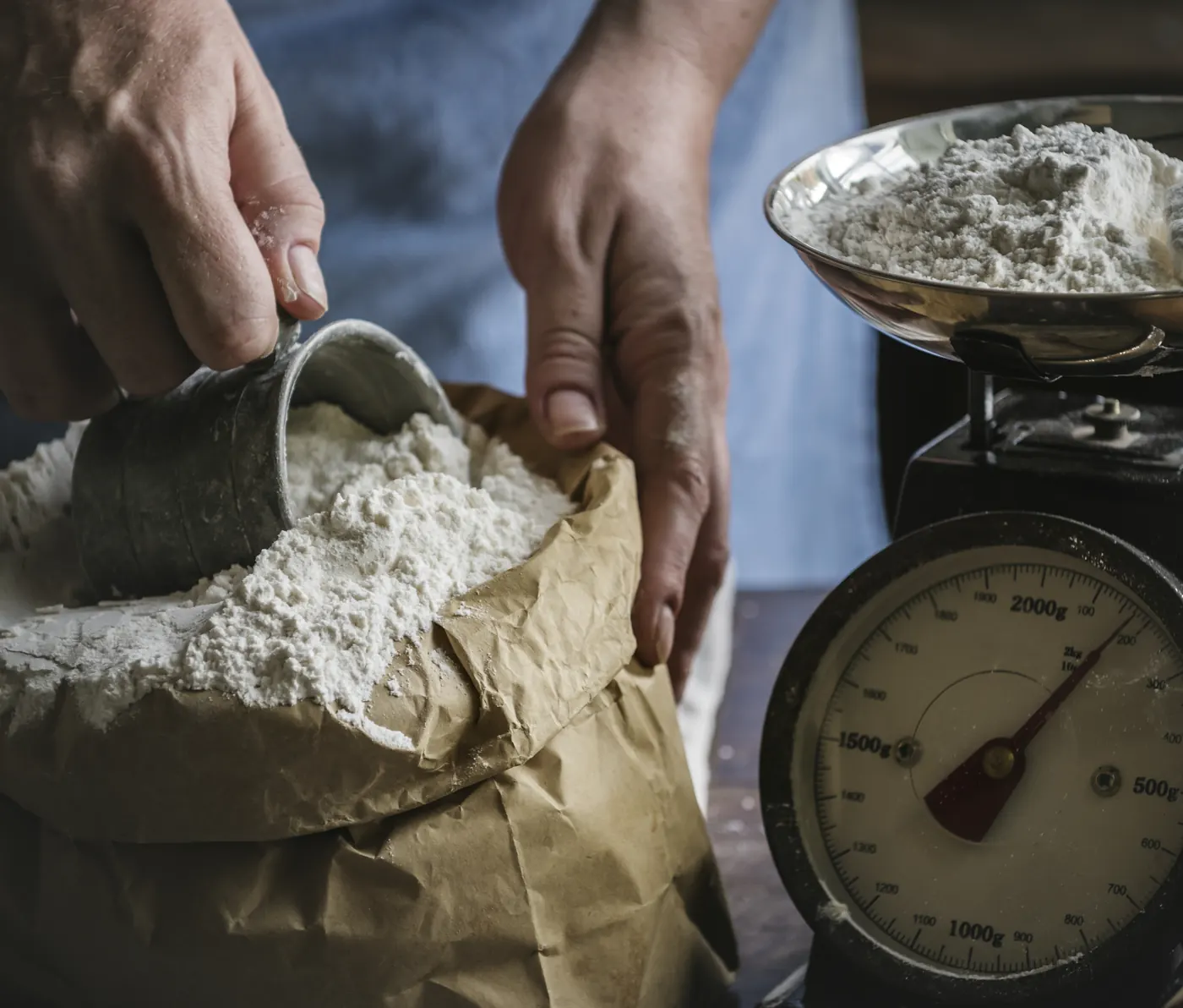 Hands using a metal cup to scoop flour from a paper bag beside a mechanical kitchen scale.