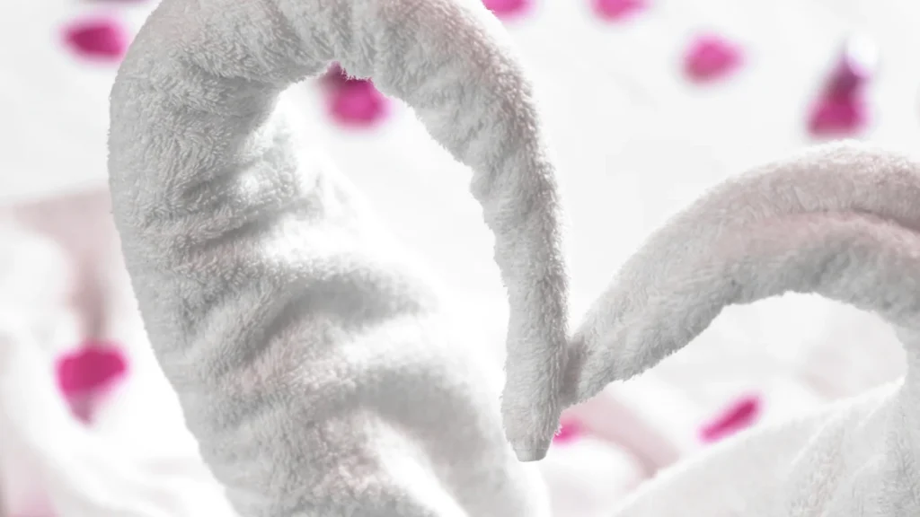 White towels folded into swan necks form a heart above scattered pink petals.