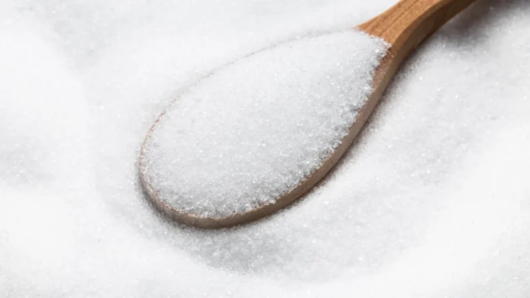 Wooden spoon filled with white sugar crystals resting on a bed of sugar