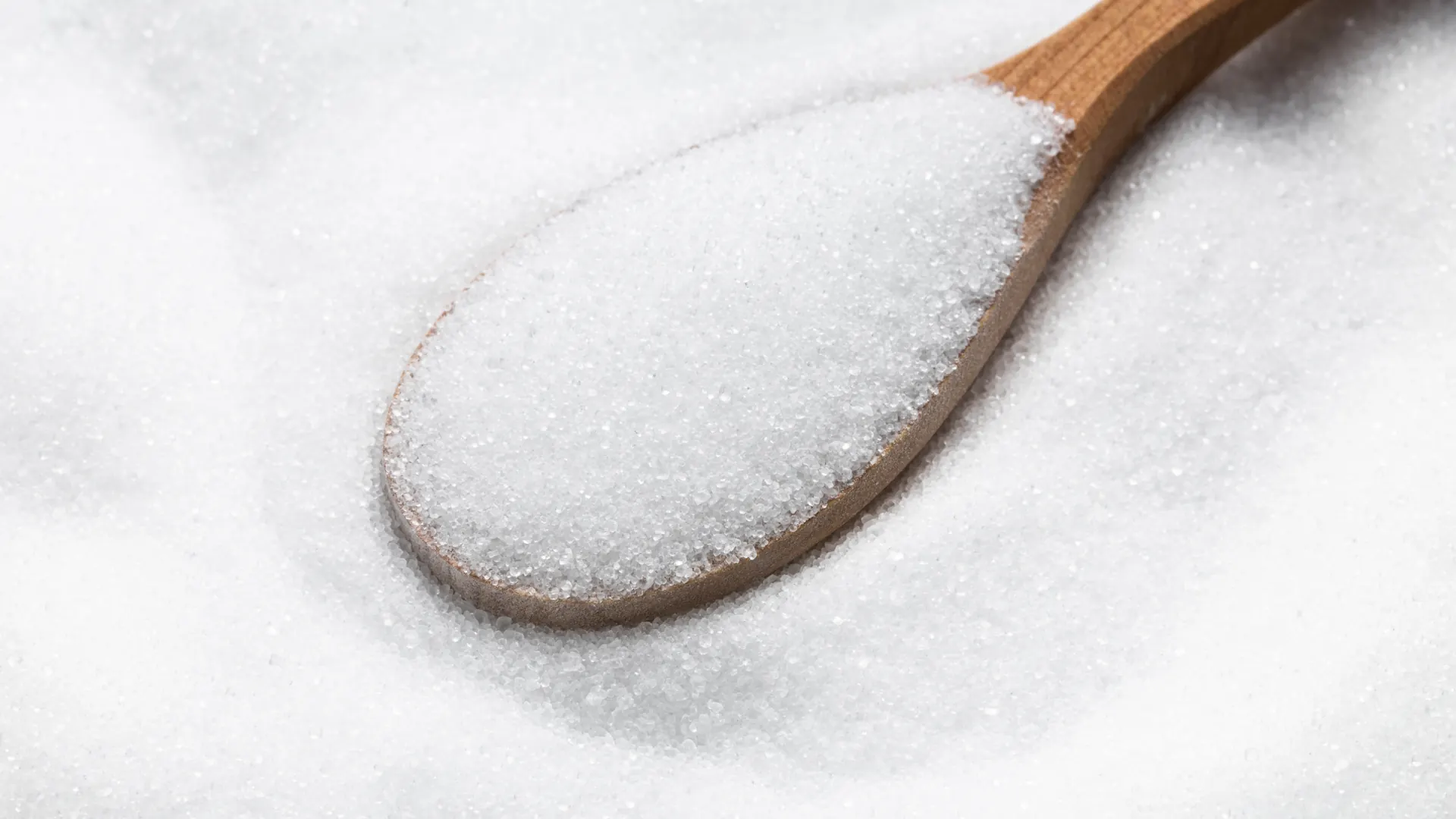 Wooden spoon filled with white sugar crystals resting on a bed of sugar