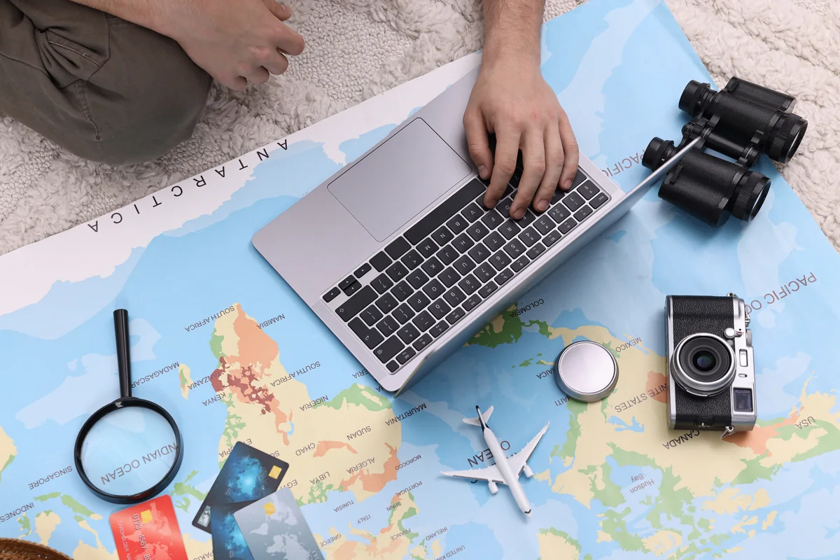 Hands using a laptop on a world map beside a camera, binoculars, passports, and travel accessories.