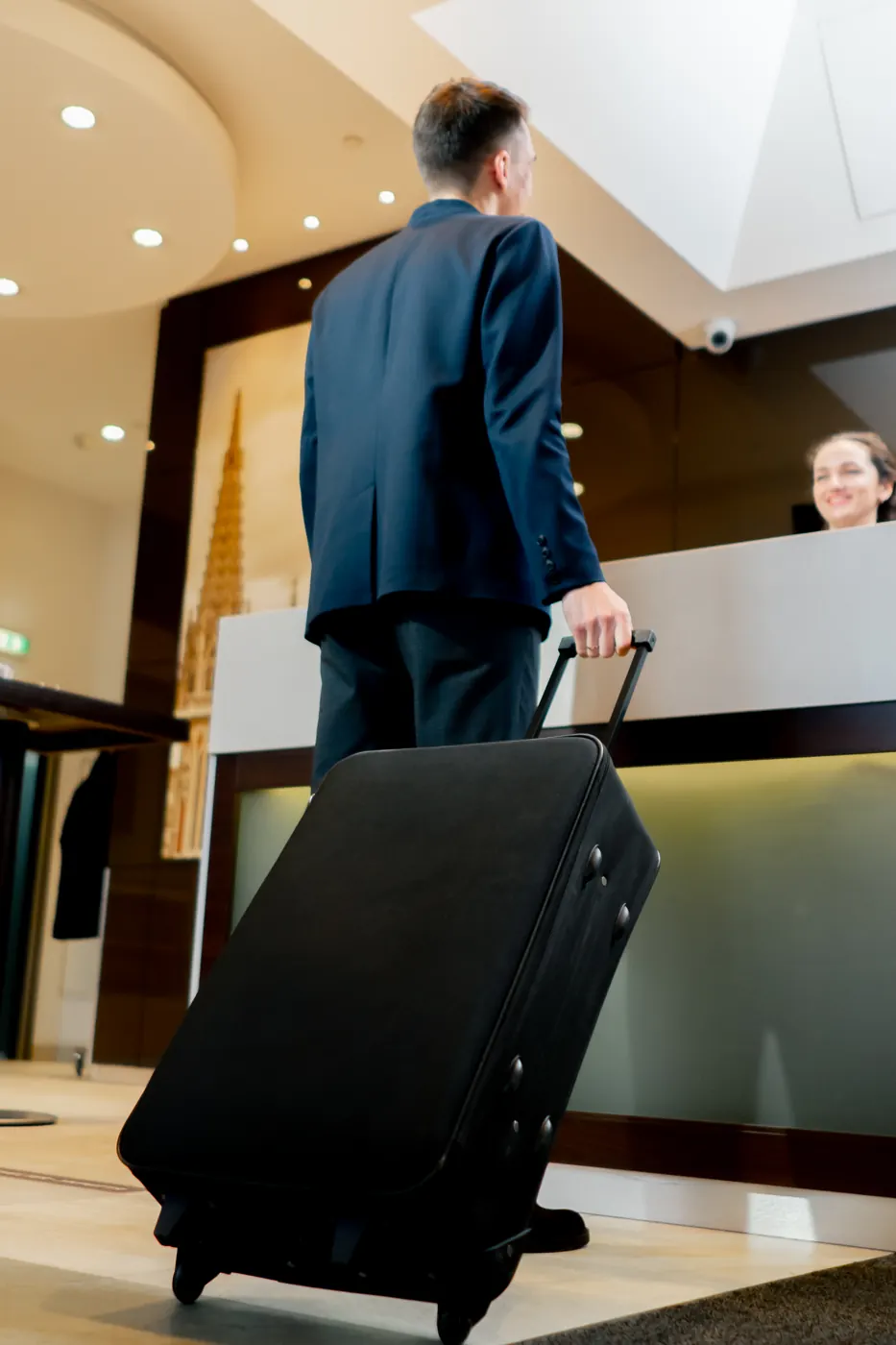 A traveler with a rolling suitcase standing at a hotel reception desk.
