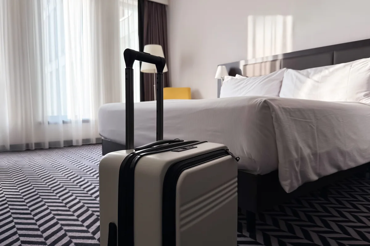 A wheeled suitcase standing beside a hotel bed near a bright window.