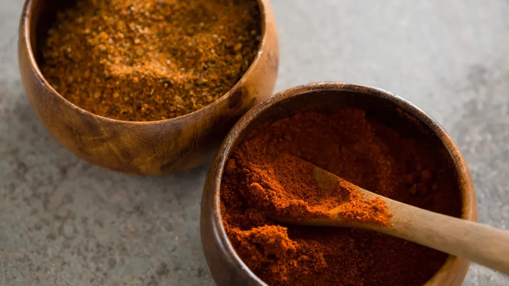 Two wooden bowls hold ground and blended spices with a wooden spoon on a gray countertop.