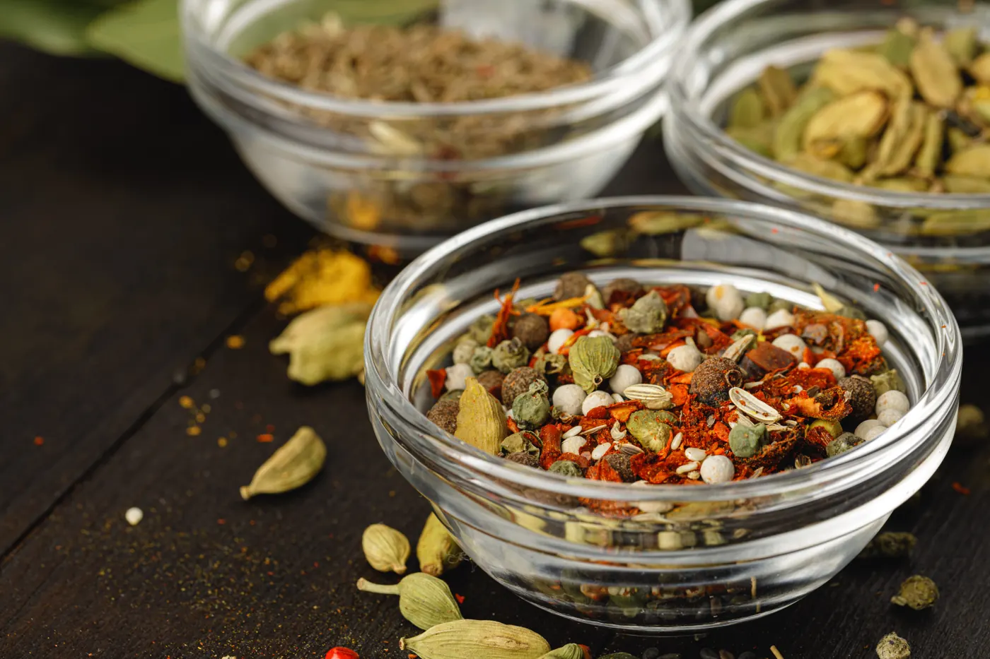 Clear glass bowls hold whole pods, mixed spices, and ground spices on a dark wooden surface.