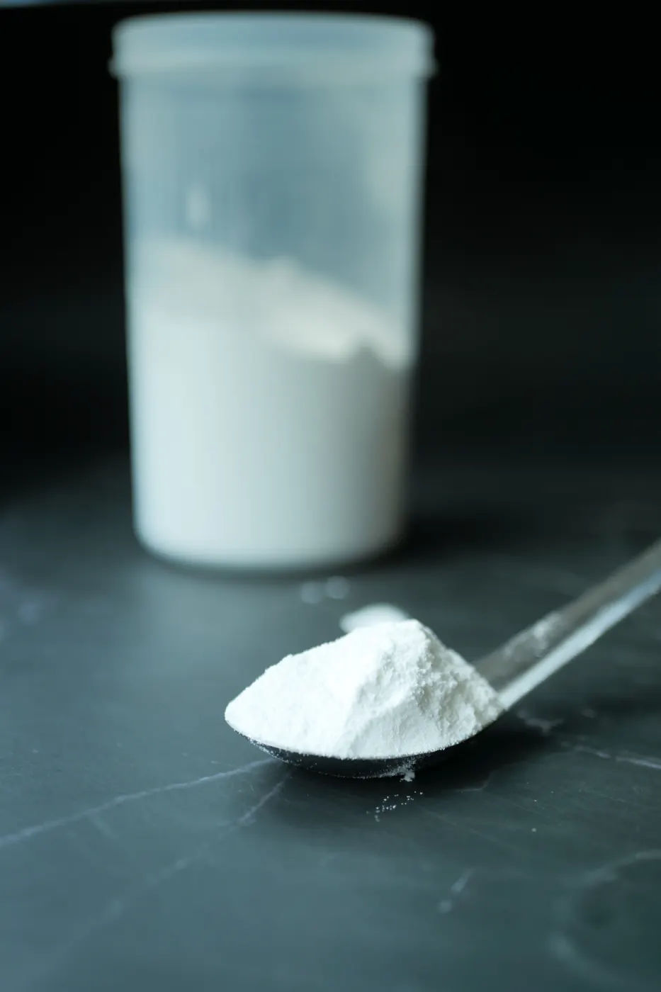 A spoon holds white powder on a dark counter, with an unmarked container behind it.