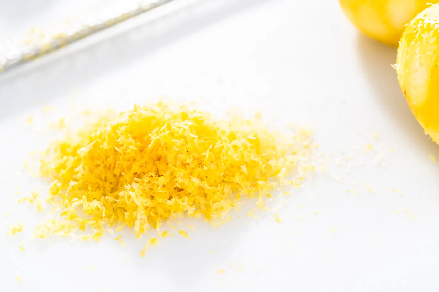 A small mound of freshly grated yellow lemon zest rests on a white cutting board beside lemons.