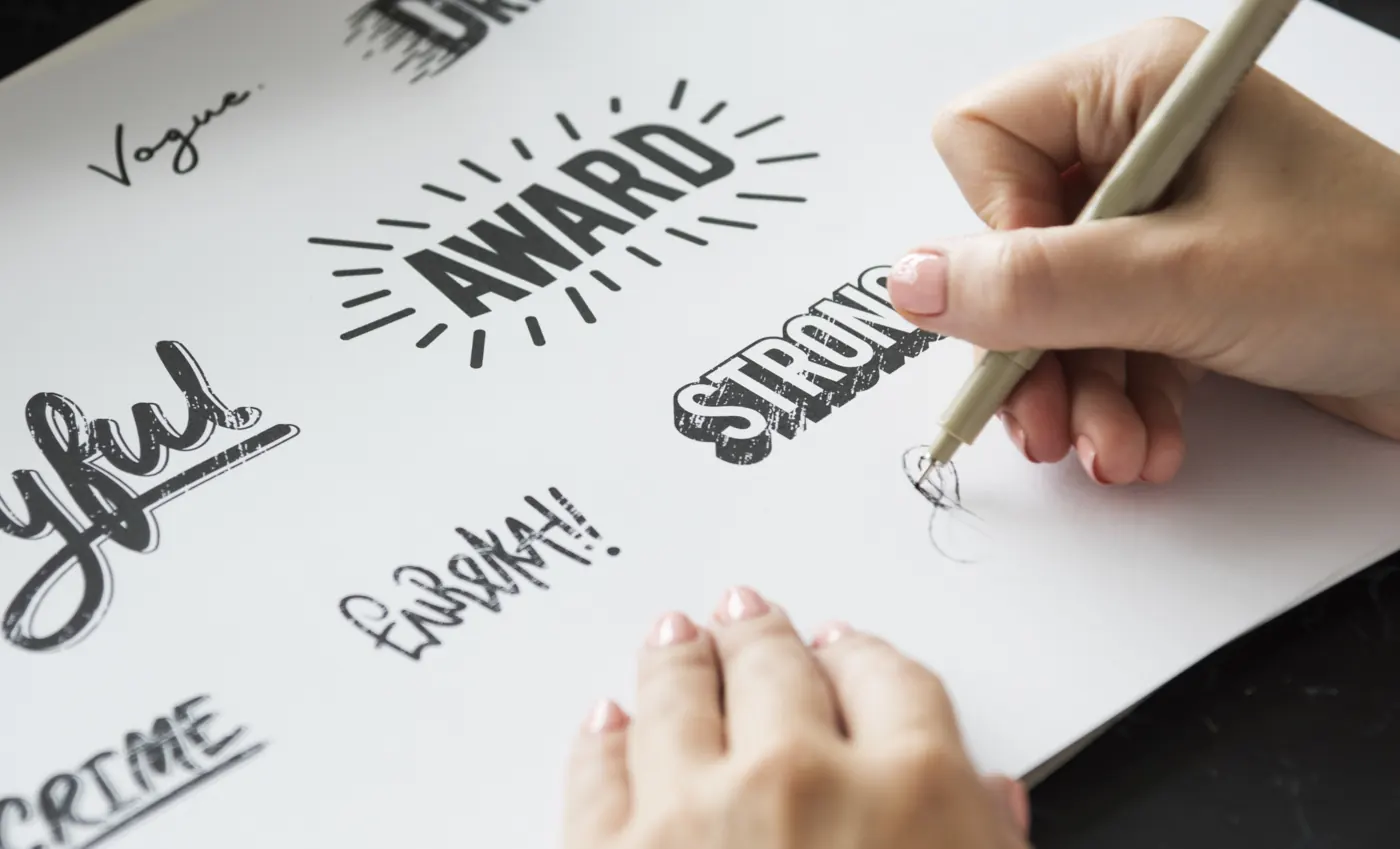 Hands drawing bold and script lettering designs on white paper.