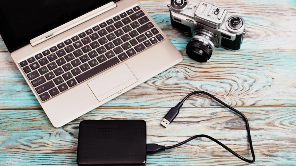Laptop, camera, and external hard drive with a USB cable arranged on a wooden desk
