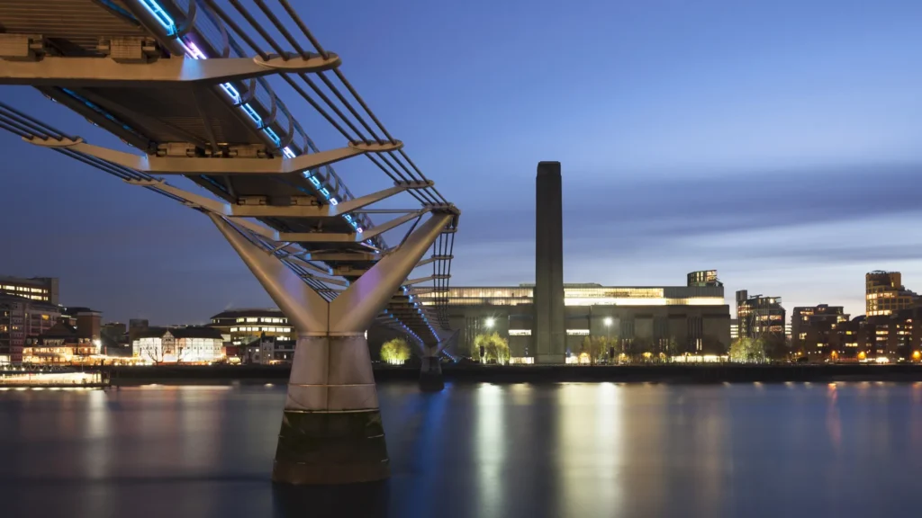 The illuminated Millennium Bridge crosses the Thames toward Tate Modern at twilight in London.