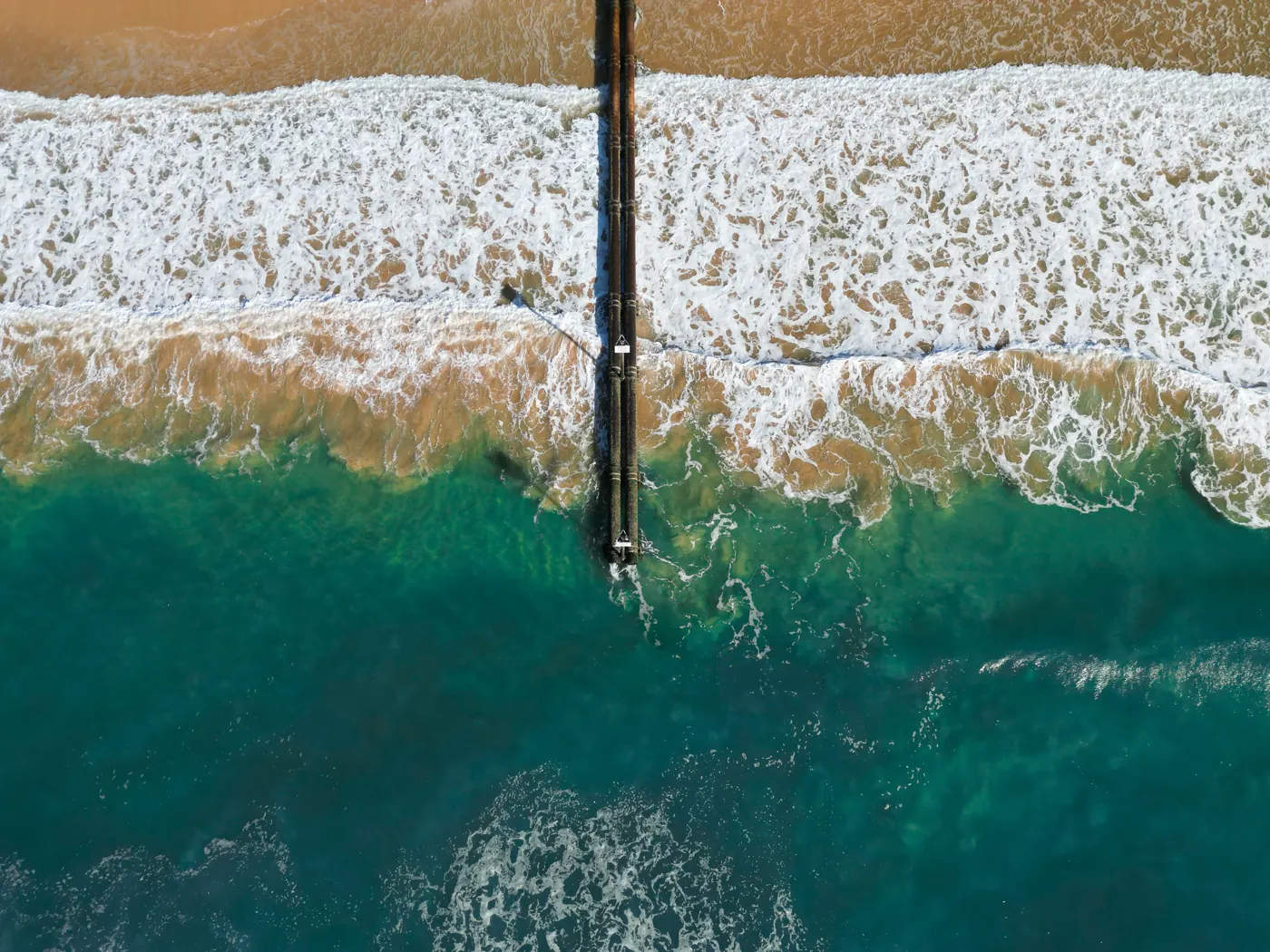 Overhead view of white surf meeting Manly Beach, with dark stormwater infrastructure extending into the water