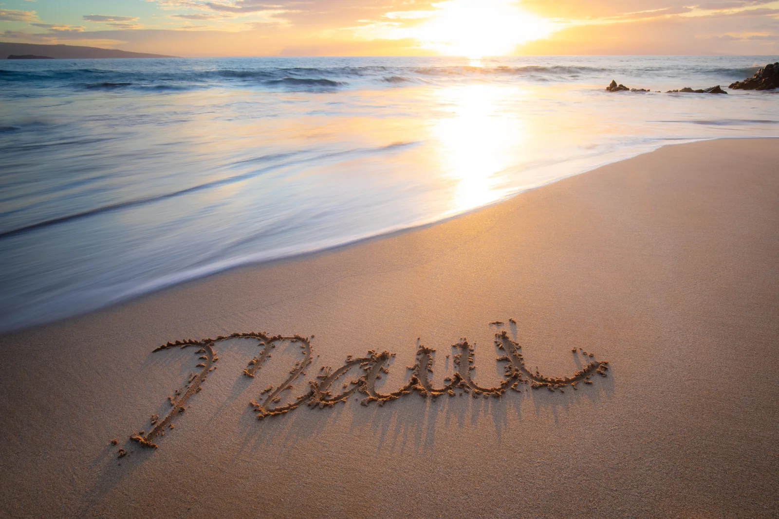 The word Maui is written in sand beside gentle surf and a setting sun.