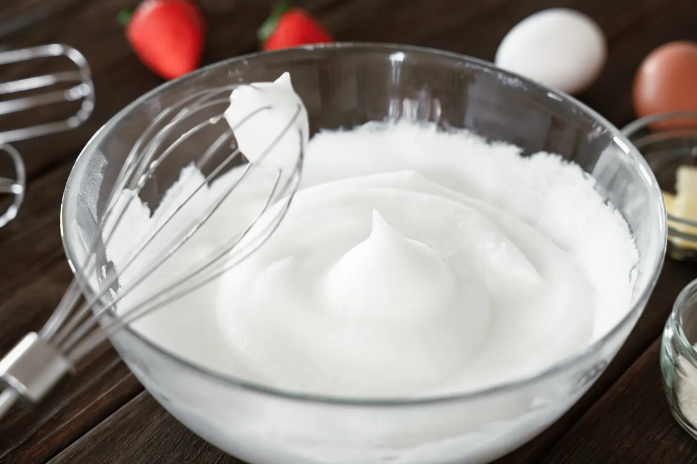 A whisk rests in whipped egg whites in a glass bowl on a wooden table.
